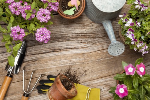 A wooden garden table with a weathered, textured surface features various gardening tools and supplies arranged in a neat manner. In the top left, a small potted plant with vibrant pink flowers rests beside a metal hand rake with a wooden handle. Towards the bottom left, a pair of pruning shears with black and silver blades is placed near a terracotta pot filled with soil, positioned above a pair of yellow gardening gloves. In the top right, a grey metal watering can with a perforated spout is angled slightly, ready for use. Surrounding the tools and pots are multiple flower pots containing blooming plants with purple and pink flowers, adding natural colour to the scene. The background includes parts of additional potted plants, indicative of a well-maintained garden workspace, likely situated outdoors or on a patio in the Docklands area. The lighting suggests a bright, possibly sunny day, highlighting the natural tones of the wood and plants. The image depicts a tidy, organized gardening setup, suitable for gardening and landscaping services offered by Hedge Trimming Docklands, reinforcing the focus on planting, pruning, and outdoor maintenance in a typical outdoor garden environment near the Docklands area in London.