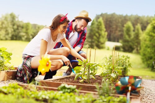 A photograph showing a woman and a man in a well-maintained garden, with the woman wearing a white top and plaid skirt, and the man dressed casually with a straw hat and a plaid shirt. They are tending to a raised garden bed filled with small green plants, possibly vegetables or herbs, with gardening tools nearby. The garden is surrounded by lush greenery, including a neatly trimmed lawn, mature trees in the background, and bordered with gravel pathways. The scene is lit by bright, natural sunlight, suggesting a clear day in the outdoor space of a residential garden or communal outdoor area, consistent with gardening services provided in the Docklands area, as explained on the website for Hedge Trimming Docklands.