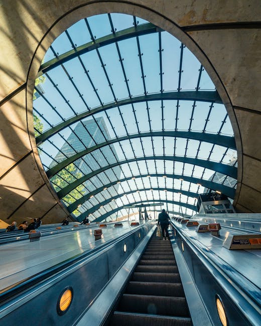 View from the bottom of an escalator inside Canary Wharf station, showing a large arched glass roof composed of multiple curved panels with metal framing, allowing natural daylight to illuminate the s