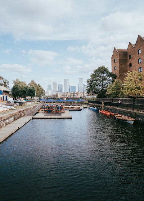 A tranquil scene along a narrow canal in an urban setting, with calm water reflecting the overcast sky. On the left side, a stone embankment features a small dock area with stacked wooden logs and a m